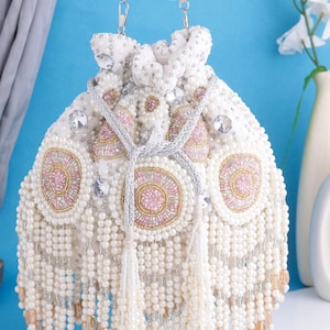 May include: A white beaded handbag with a drawstring closure and a silver chain strap. The bag features pink and gold circular embellishments and dangling pearl strands. A decorative and elegant accessory.
