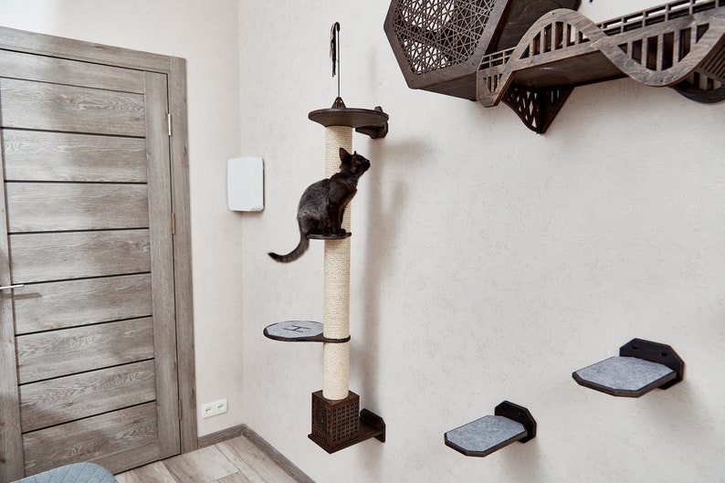 May include: A black cat climbing a tall, beige scratching post with a brown platform. The post is mounted on a white wall with two smaller platforms below. The platforms are covered in gray fabric.