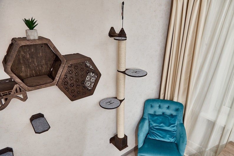 May include: A beige cat tree with three platforms and a scratching post. The platforms are covered in gray felt and the post is wrapped in sisal rope. The cat tree is mounted to a wall and is next to a teal armchair with a teal cushion. The cat tree is in a room with white walls and beige curtains.