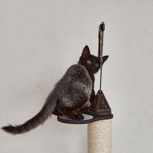 May include: A black kitten with green eyes sits on a cat tree, looking over its shoulder. The cat tree has a brown platform and a beige rope-covered post.