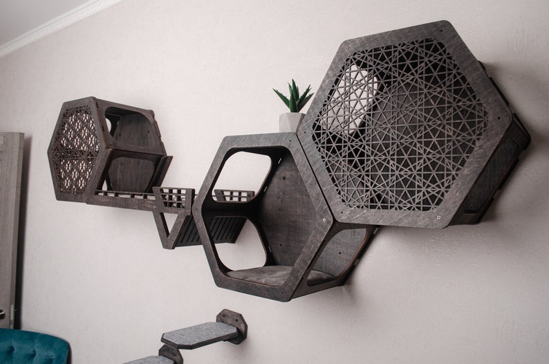 Puede incluir: Un conjunto de tres estantes hexagonales para gatos hechos de madera oscura con un patr&oacute;n geom&eacute;trico. Los estantes est&aacute;n fijados a la pared y tienen una peque&ntilde;a plataforma para que los gatos puedan saltar.