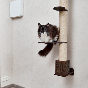 May include: A black and white cat sits on a platform of a tall, beige, sisal-covered cat scratching post. The post is mounted to a white wall.