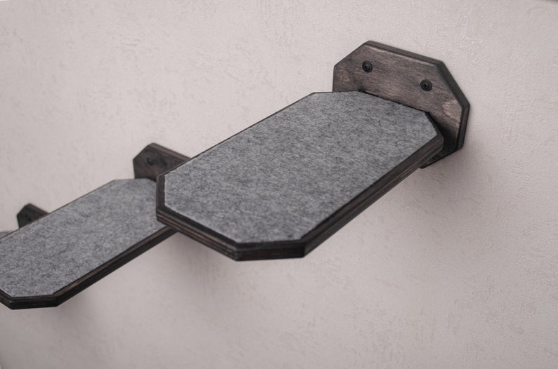 Puede incluir: Dos estantes grises para gatos con marcos de madera negra montados en una pared blanca. Los estantes est&aacute;n inclinados hacia arriba y tienen una superficie suave y texturizada.
