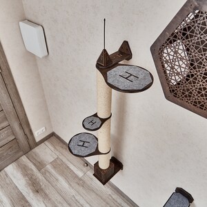 May include: A wall-mounted cat tree with a sisal scratching post and three platforms covered in gray fabric. The platforms are shaped like rounded rectangles and have the letter "H" printed on them. The cat tree is mounted on a white wall.