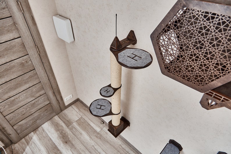May include: A wall-mounted cat tree with a sisal scratching post and three platforms covered in gray fabric. The platforms are shaped like rounded rectangles and have the letter "H" printed on them. The cat tree is mounted on a white wall.