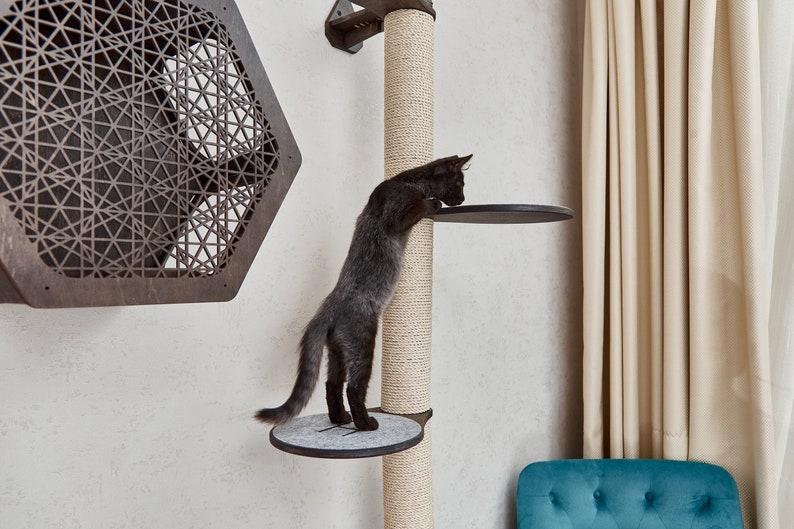 May include: A black cat climbing a sisal-covered scratching post with two platforms. The post is mounted to a white wall. The cat is looking down at the lower platform.