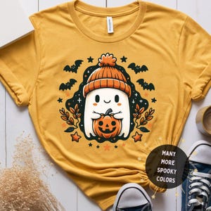 May include: Mustard yellow t-shirt featuring a cartoon ghost wearing an orange beanie and holding a jack-o'-lantern. The design includes bats, stars, and foliage. The shirt has the text "MANY MORE SPOOKY COLORS" in a circular graphic.