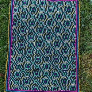 May include: A colourful woven rug with a geometric pattern in shades of green, yellow, pink, and white. The rug has a black background and a border of yellow, pink, and white.