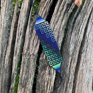 May include: A beaded bracelet with a geometric pattern in shades of blue, green, and black. The bracelet is displayed on a weathered wooden surface, showcasing its intricate design and color gradient.