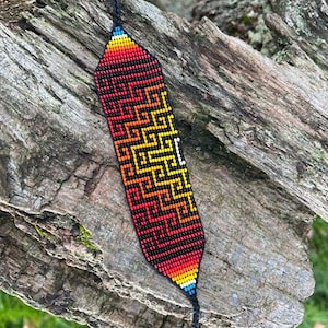 May include: A beaded bracelet with a geometric pattern in red, orange, yellow, and black. The bracelet has a rectangular shape and a black cord fastening. The design features a repeating zigzag pattern.