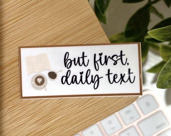 May include: A rectangular sticker with a brown border and the text "but first, daily text" in black script. The sticker also features a small open book, a coffee cup with a heart, and a coffee bean illustration.