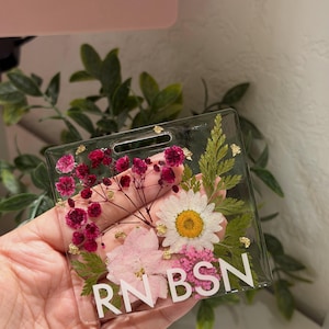 May include: A square clear resin badge reel featuring embedded pressed flowers including a white daisy, pink petals, and dark pink baby's breath, accented with gold leaf flakes. The white text RN BSN is printed across the bottom of the piece.