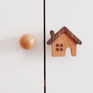 May include: A wooden house-shaped cabinet knob with a brown wood roof and a light brown wood body. The knob has a small window and a door. The knob is attached to a white surface.