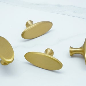 May include: Four gold oval cabinet knobs with a brushed finish. Two of the knobs have a simple oval shape, while the other two have a more elaborate design with a raised base.