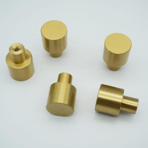 May include: Five brass cabinet knobs with a cylindrical design. The knobs have a smooth, matte finish and a simple, modern aesthetic. The knobs are in various orientations, showcasing their shape and construction.