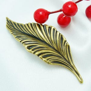 May include: A gold-toned metal leaf brooch with a detailed, textured design. The brooch is on a white background.