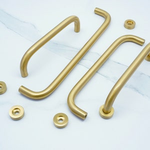 May include: Four gold-toned cabinet pulls with rounded edges. The pulls are arranged in a staggered pattern on a white marble surface.