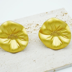 May include: Two gold-colored flower-shaped earrings. Each earring features a detailed center and petal design. The earrings are displayed on a textured, light-colored surface, creating a contrast with the bright gold.