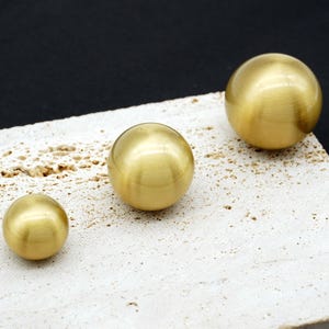 Peut inclure: Trois boutons de meuble sphériques en laiton de tailles différentes reposent sur une surface texturée blanc cassé sur fond noir. Les sphères ont une finition métallique lisse, reflétant la lumière. Ces boutons sont décoratifs.