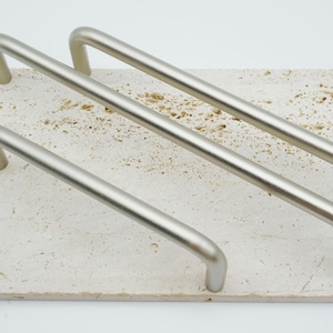 以下が含まれることがあります： 長方形の明るい色の石タイルに配置された、3つのつや消しニッケルキャビネットプル。プルはシンプルで湾曲したデザインで、石は小さな穴のあるテクスチャード加工された表面を持っています。