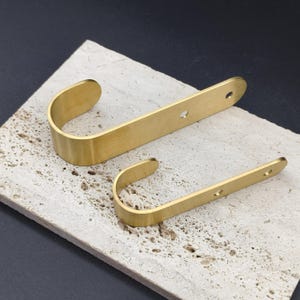 May include: Two brass-colored metal hooks. Each hook has a curved, U-shaped design with a flat, rectangular mounting plate. The hooks are on a textured, light-colored surface, with a dark background.