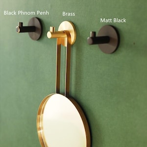 May include: Three wall hooks in black and brass are shown against a green backdrop. The hooks are labelled "Black Phnom Penh", "Brass", and "Matt Black". A round brass mirror hangs from the brass hook.