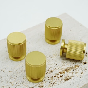 May include: Four gold-colored cabinet knobs with a cylindrical design and ridged texture. The knobs have a flat top and a small base, and are arranged on a light-colored, textured surface.