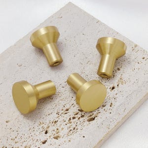 Könnte beinhalten: Vier Messing-Schrankknöpfe mit goldener Oberfläche sind auf einer strukturierten, hellfarbigen Oberfläche angeordnet. Zwei Knöpfe haben eine konische Form, während die anderen beiden eine flache, kreisförmige Fläche haben. Diese Beschläge sind ideal, um Möbeln einen Hauch von Eleganz zu verleihen.