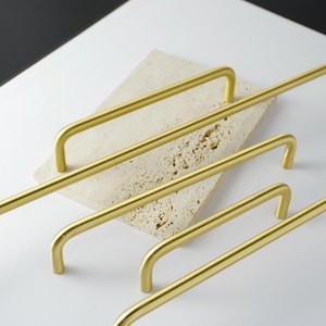 May include: A collection of gold-colored, rectangular cabinet pulls with a curved handle design. The pulls are arranged on a light-colored, textured surface, with a contrasting dark background. The hardware is likely for kitchen or bathroom cabinets.