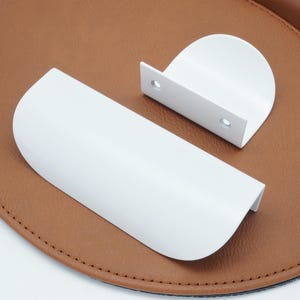 Peut inclure: Deux poignées de meuble en métal blanc. L'une est une poignée rectangulaire incurvée et l'autre est une poignée rectangulaire arrondie.
