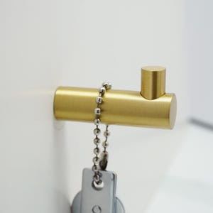 May include: A gold metal hook with a round base and a chain attached. The hook is mounted on a white wall.