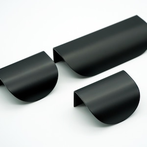 Può includere: Tre maniglie per mobili nere opache di diverse dimensioni. Le maniglie hanno un design curvo a mezzaluna con un'estetica semplice e moderna. Le maniglie sono in metallo e sono poste su uno sfondo bianco.