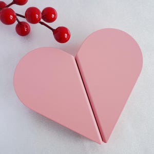 Puede incluir: Un objeto de color rosa en forma de corazón hecho de dos piezas separadas que encajan. El corazón está sobre un fondo blanco.