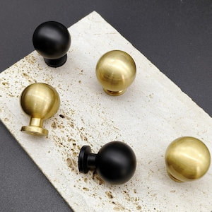 Puede incluir: Un surtido de pomos de armario en acabados negro mate y latón cepillado. Los pomos son esféricos con una base cilíndrica corta. Los pomos están dispuestos sobre una superficie texturizada de color claro sobre un fondo oscuro.