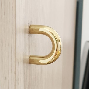 Puede incluir: Un tirador de armario dorado en forma de D montado sobre una superficie de madera clara. El tirador tiene un acabado liso y reflectante, y la veta de la madera es visible. El tirador tiene un diseño de semicírculo y la ferretería está al ras de la madera.