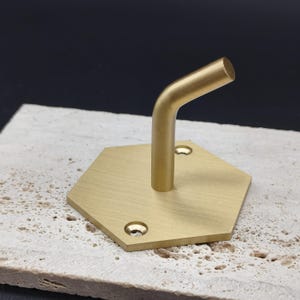 Może przedstawiać: Mosiężny, sześciokątny wieszak ścienny z zakrzywionym ramieniem. Hak ma prosty, nowoczesny design z dwoma otworami na śruby do montażu. Hak jest wykonany z metalu i ma matowe wykończenie.