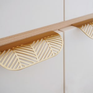 May include: Two gold-toned cabinet pulls with a leaf-shaped design and a geometric pattern. The pulls are made of metal and have a brushed finish.