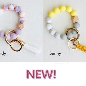 May include: Two silicone beaded keychains with a tassel. The keychain on the left has white, purple, and pink beads with a wooden bead. The keychain on the right has yellow, white, and gray beads with a wooden bead. Both keychains have a gold key ring.