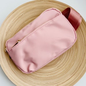 K&ouml;nnte beinhalten: Eine rosa Rei&szlig;verschlusstasche mit einem goldenen Rei&szlig;verschluss und einem rosa Riemen. Die Tasche liegt auf einer Holzoberfl&auml;che.