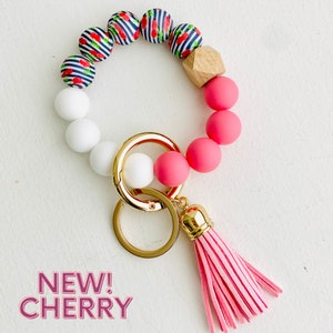 May include: A pink and white beaded keychain with a cherry print, a gold key ring, and a pink tassel. "NEW! CHERRY" is written in black text on a white background.