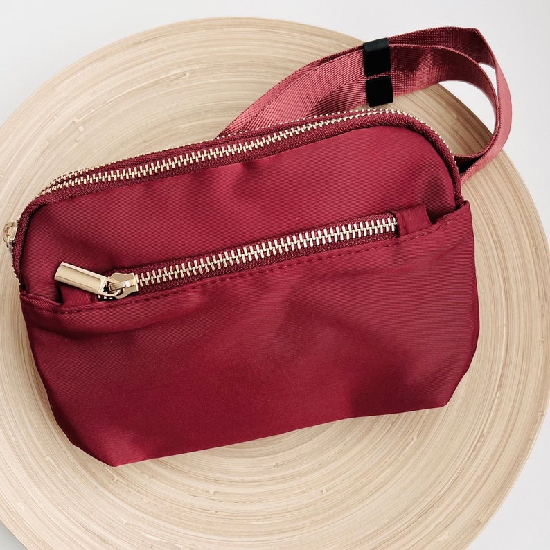 K&ouml;nnte beinhalten: Eine bordeauxfarbene Bauchtasche mit einem goldenen Rei&szlig;verschluss und einem schwarzen verstellbaren Riemen.