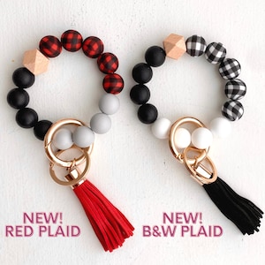 May include: Two silicone beaded keychains with a tassel. One keychain has red and black plaid beads, a wooden bead, and a red tassel. The other keychain has black and white plaid beads, a wooden bead, and a black tassel. Both keychains have black silicone beads and a gold key ring.