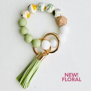 May include: A green and white beaded keychain with a floral pattern and a green tassel. The keychain has a gold key ring and a wooden bead.