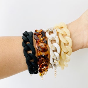 May include: Four chunky chain bracelets stacked on a wrist. The bracelets are black, brown tortoise shell, white with brown marbling, and cream.