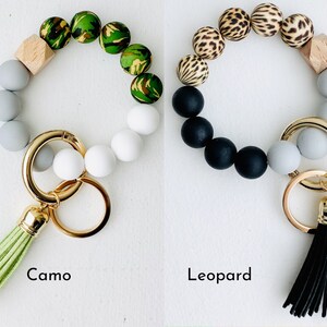 May include: Two beaded keychains with a wooden bead and a tassel. One keychain has camouflage beads and a green tassel. The other keychain has leopard print beads and a black tassel. The text "Camo" is below the first keychain and "Leopard" is below the second keychain.