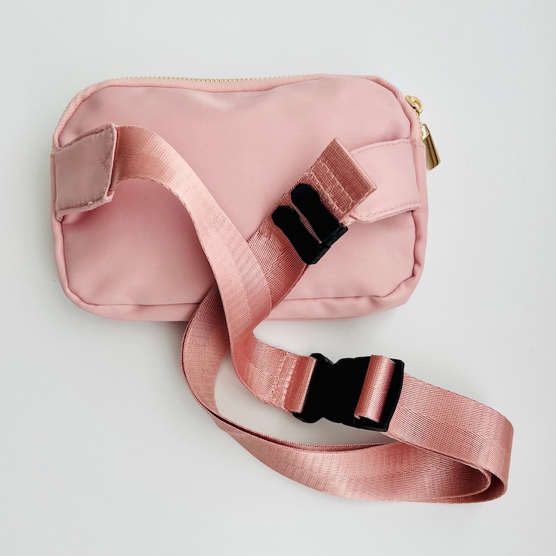 K&ouml;nnte beinhalten: Eine rosa Bauchtasche mit einem verstellbaren schwarzen Gurt und einem goldenen Rei&szlig;verschluss. Der Gurt hat zwei schwarze Schnallen.