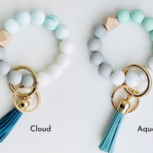 May include: Two silicone beaded keychains with a gold key ring and tassel. The keychain on the left is white, gray, and light blue with a wooden bead. The keychain on the right is gray, white, and light green with a wooden bead. The keychains are labeled "Cloud" and "Aqua".