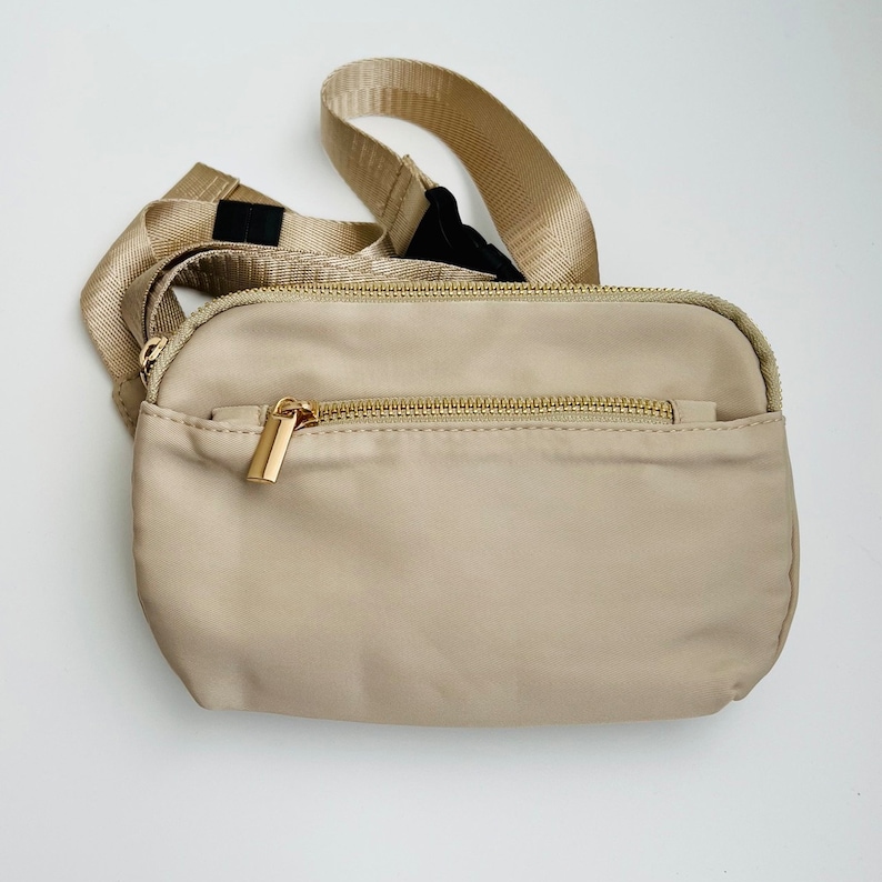 K&ouml;nnte beinhalten: Eine beige Bauchtasche mit einem goldenen Rei&szlig;verschluss und einem schwarzen verstellbaren Riemen.