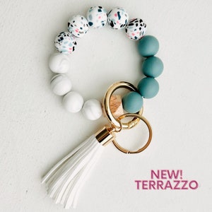 May include: A white and teal beaded keychain with a white tassel. The beads are made of silicone and have a terrazzo pattern. The keychain has a gold key ring.