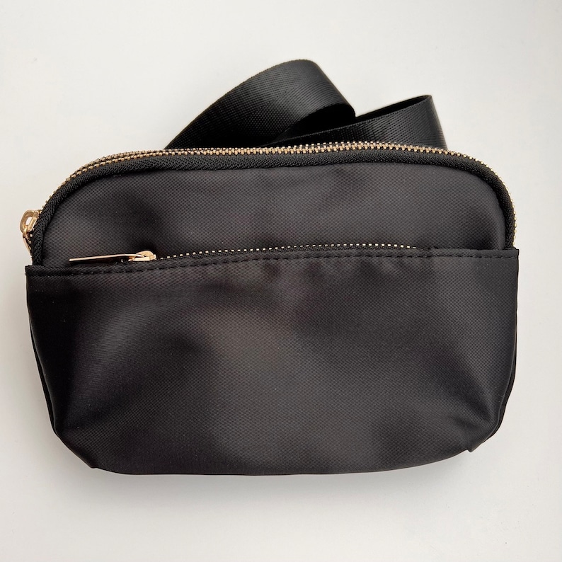 K&ouml;nnte beinhalten: Eine schwarze Nylon-Umh&auml;ngetasche mit einem goldenen Rei&szlig;verschluss und einem schwarzen verstellbaren Riemen.